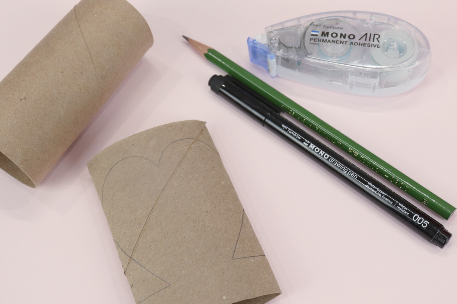 Image contains two empty toilet paper rolls, one of which is flattened with a heart shape sketched on top. A Tombow Pencil, Tombow MONO Drawing Pen, and adhesive runner sit nearby on a light pink background.
