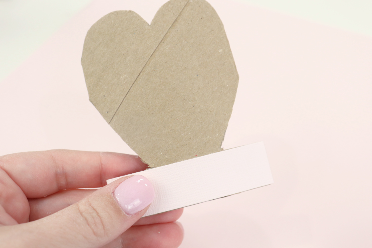 Image contains Amy’s hand holding a toilet paper roll cut into a heart shape with pink cardstock wrapped around the bottom.