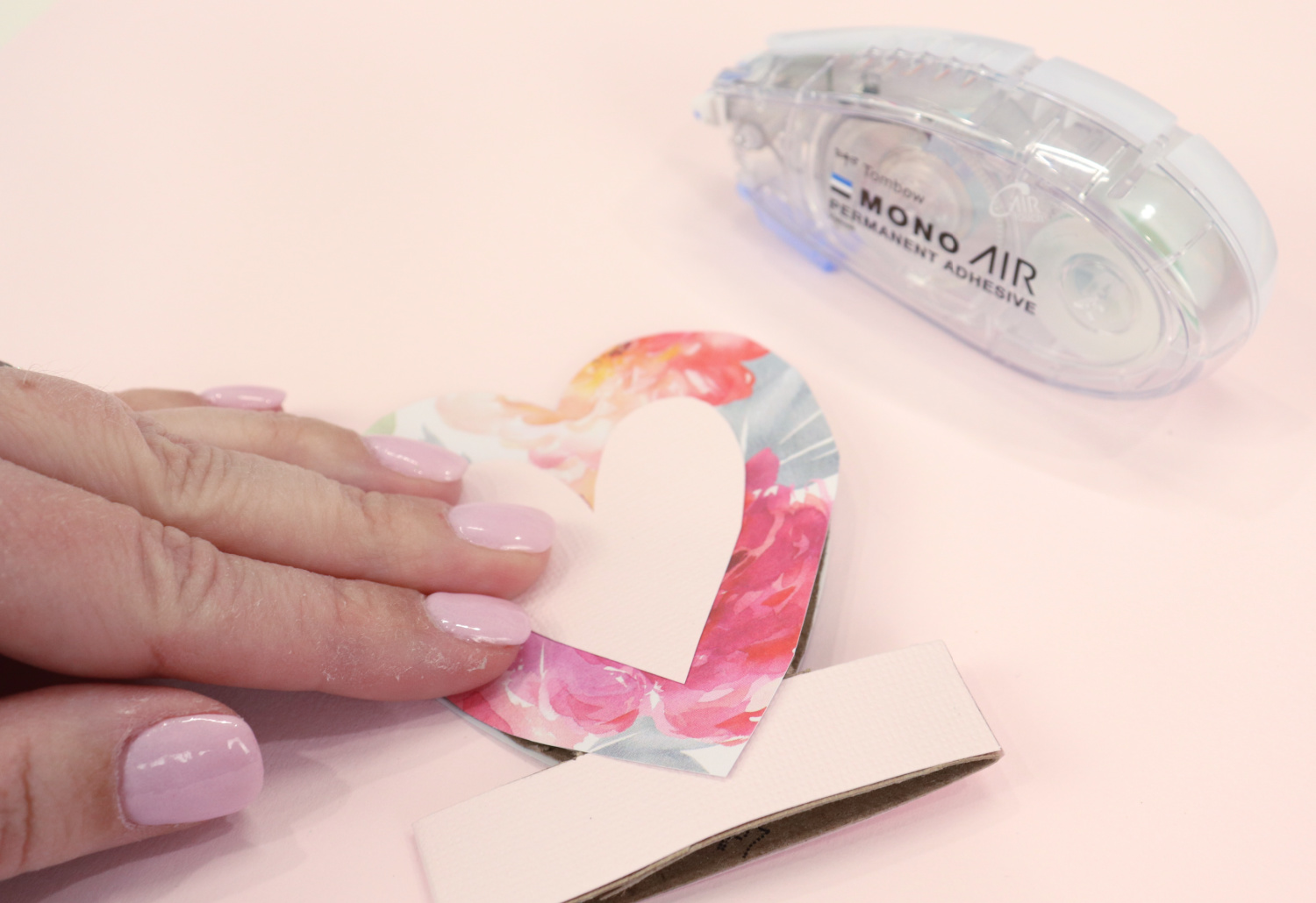 Image contains Amy’s hand pressing heart shaped scrapbook paper onto the paper roll. An adhesive runner sits off to the side on a pink background.