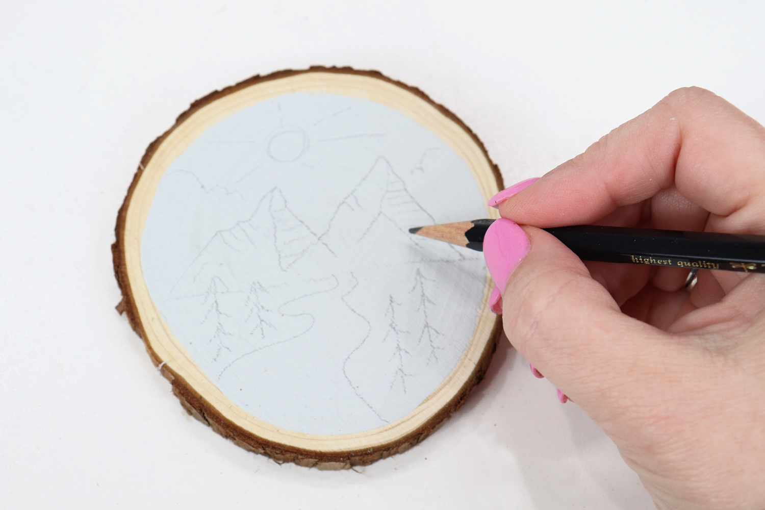 Image contains Amy’s hand holding a Tombow drawing pencil and sketching a scene on a painted wood slice.