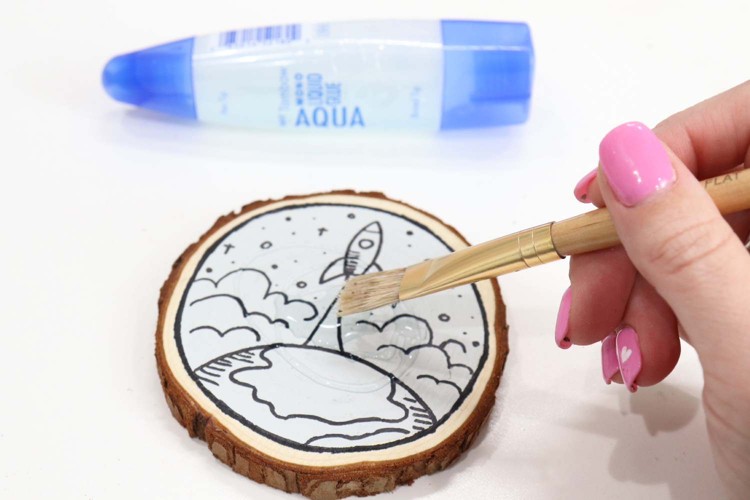 Image contains Amy’s hand holding a paintbrush and spreading MONO Aqua Liquid Glue on the surface of the wood slice coaster. The tube of glue sits nearby.