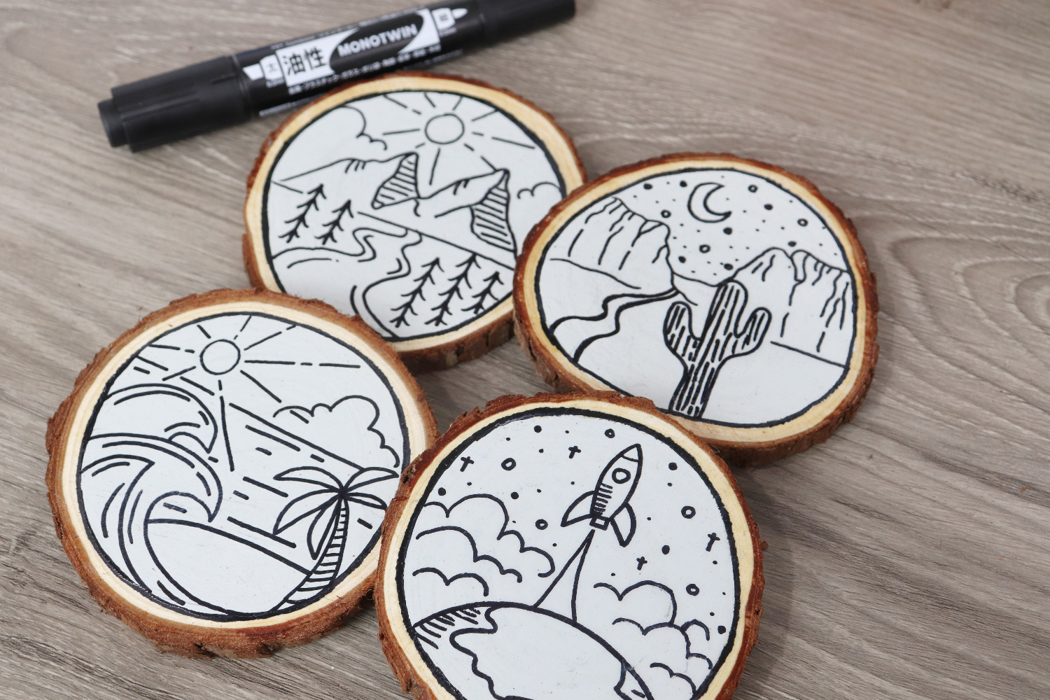 Image contains four monoline travel coasters: one is beach themed, one mountain themed, one desert themed, and one space themed, on a white background. A MONOTwin marker sits nearby on a wooden desktop.