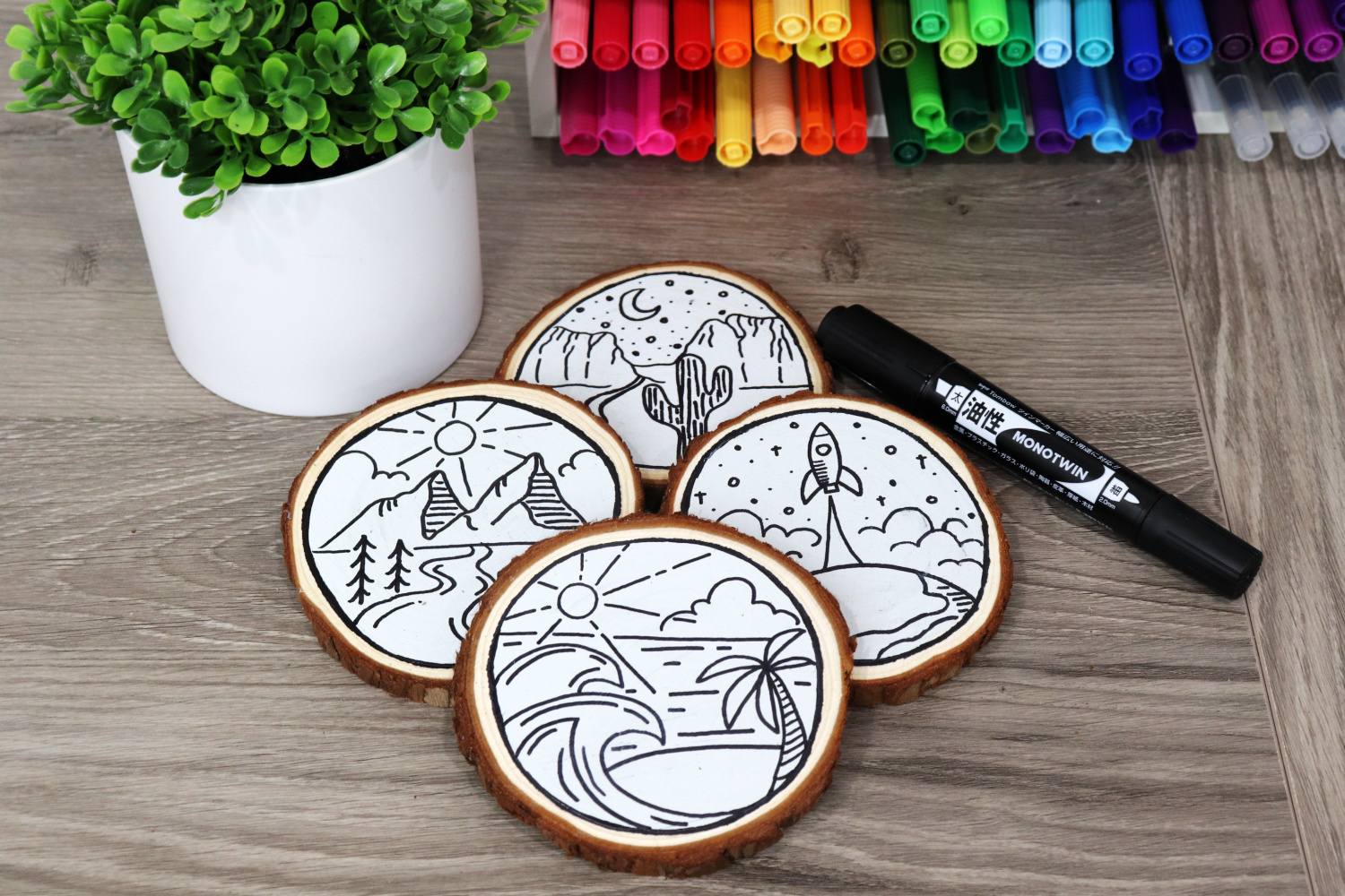 Image contains four monoline travel coasters: one is beach themed, one mountain themed, one desert themed, and one space themed, on a wooden desk. Surrounding them are a faux plant, an organizer filled with multicolored markers, and a MONOTwin bold marker.