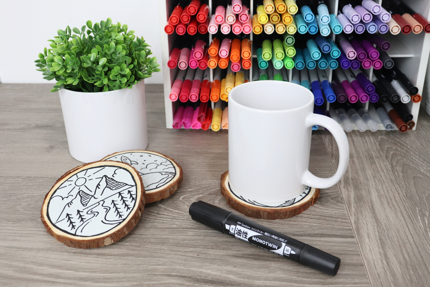 Image contains a wooden desktop with a colorful marker organizer and a faux plant, as well as a white coffee cup on a wood slice coaster. Two other monoline travel coasters sit by the plant, as does a MONOTwin bold marker.