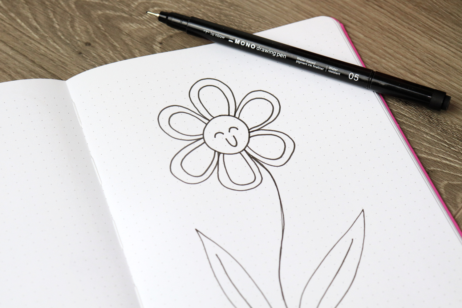 Image contains an open sketchbook on a wooden desk. A smiling flower is drawn on the page, and a MONO Drawing Pen 05 sits on top.