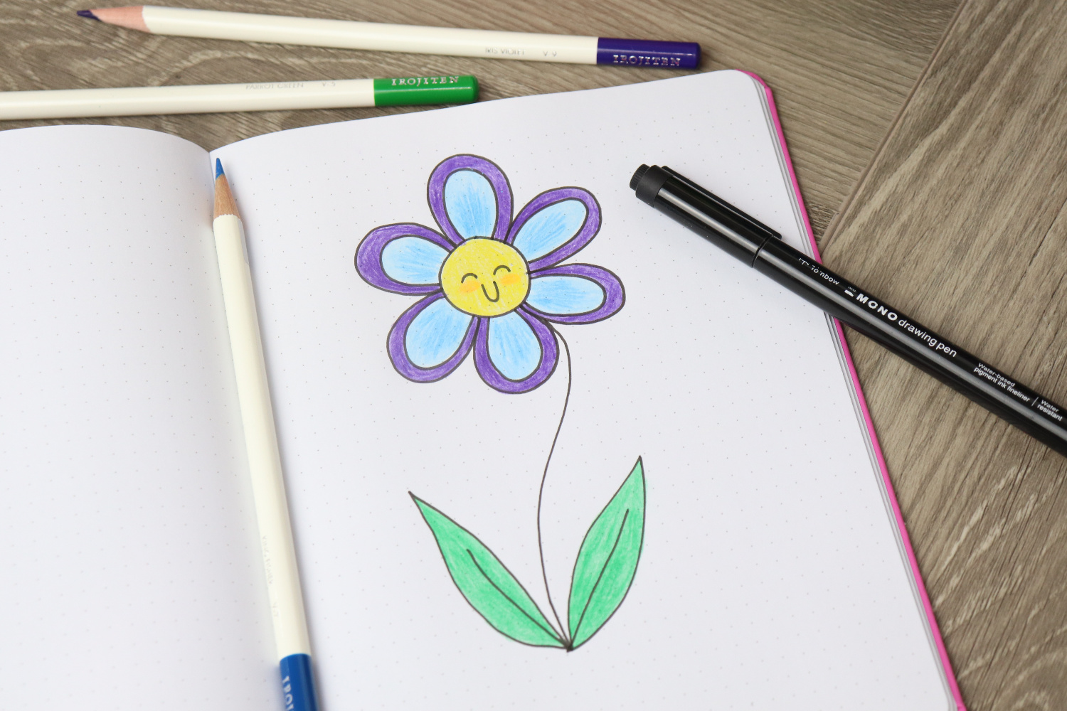 Image contains an open sketchbook on a wooden desk, with a flower doodled on the page. It is colored with purple, blue, yellow, and green colored pencils, which sit on the desk around it, along with a MONO Drawing Pen.