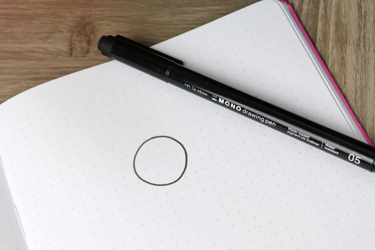Image contains an open sketchbook with a circle drawn on the page, and a Tombow MONO Drawing Pen 05 sitting on top.