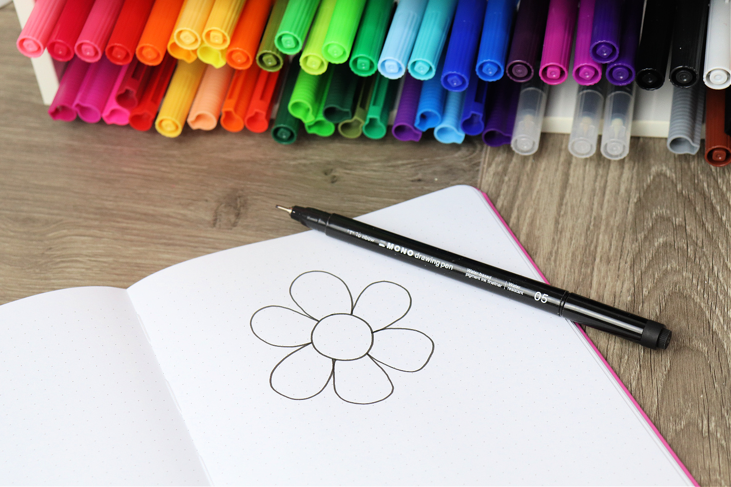 Image contains an open sketchbook sitting on a wooden desk in front of a container filled with multicolored markers. A simple flower with a round center and six petals is drawn on the page, and a MONO Drawing Pen 05 sits on top.