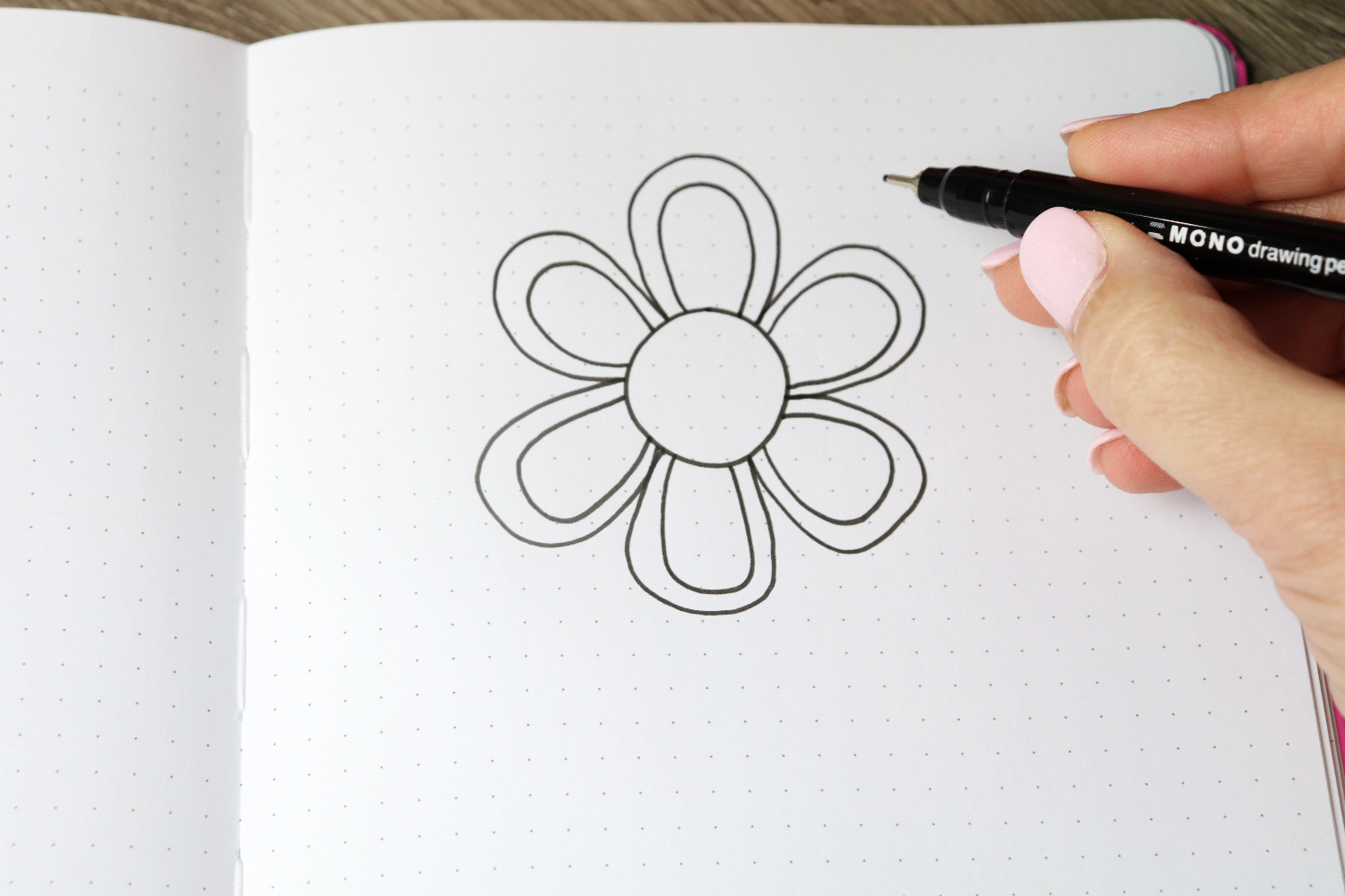 Image contains an open sketch book with a six petaled flower drawn on the right-hand page. Amy’s hand holds a MONO Drawing Pen 05 above the page.