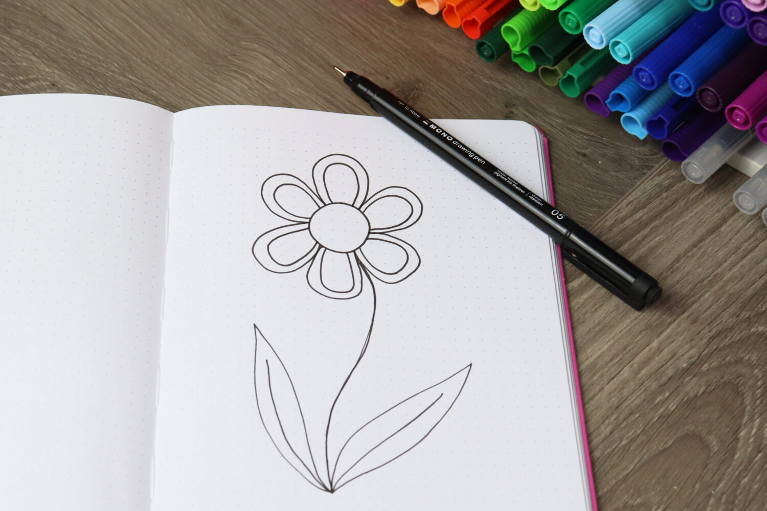 Image contains an open sketchbook next to a container of colored markers on a wooden desk. A large flower is doodled on the page, and a MONO Drawing Pen 05 sits on top.