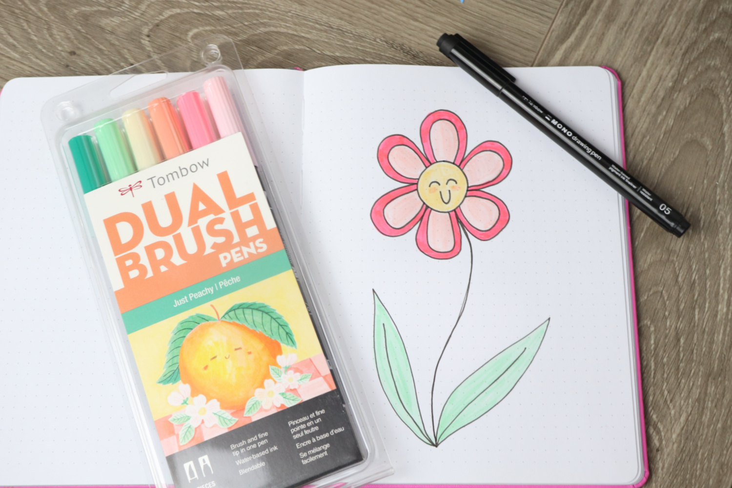 Image contains an open sketchbook on a wooden desk, with a large flower drawn on the page. It is colored pink, yellow, and green. A pack of Just Peachy Dual Brush Pens and a MONO Drawing Pen sit on top of the sketchbook.