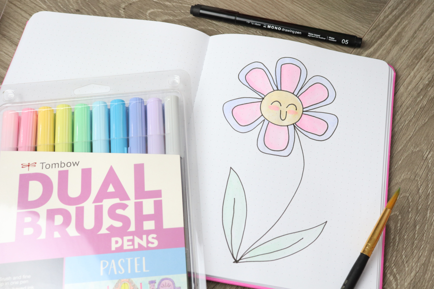 Image contains an open sketchbook on a wooden desk, with a large purple, pink, yellow, and green flower drawn on the page. A pack of pastel Dual Brush Pens, a paintbrush, and a MONO Drawing Pen sit nearby.