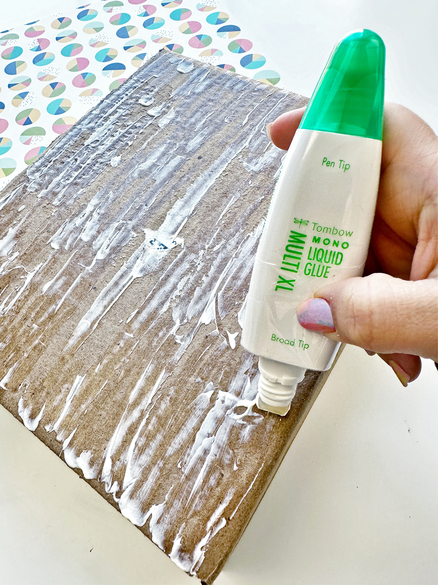 Pick a pattern paper to cover the box. Apply the Tombow MONO Multi XL Liquid Glue. Spread the glue on the cardboard using the broad tip. Once you are done, it's easy to clean. Use a paper towel to remove the excess. #tombow #recycled