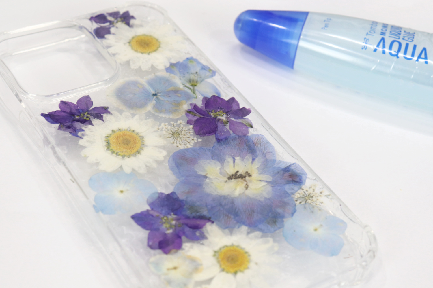 Image contains a clear phone case decorated with an assortment of white, blue, and purple pressed flowers on a white table. A tube of Tombow MONO Aqua Liquid Glue sits off to the side.