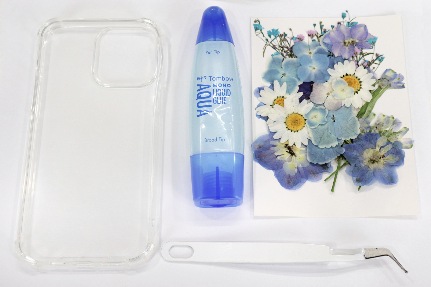 Image contains a clear phone case, a tube of Tombow MONO Aqua Liquid Glue, a pair of tweezers, and an assortment of blue and white pressed flowers on a white background.
