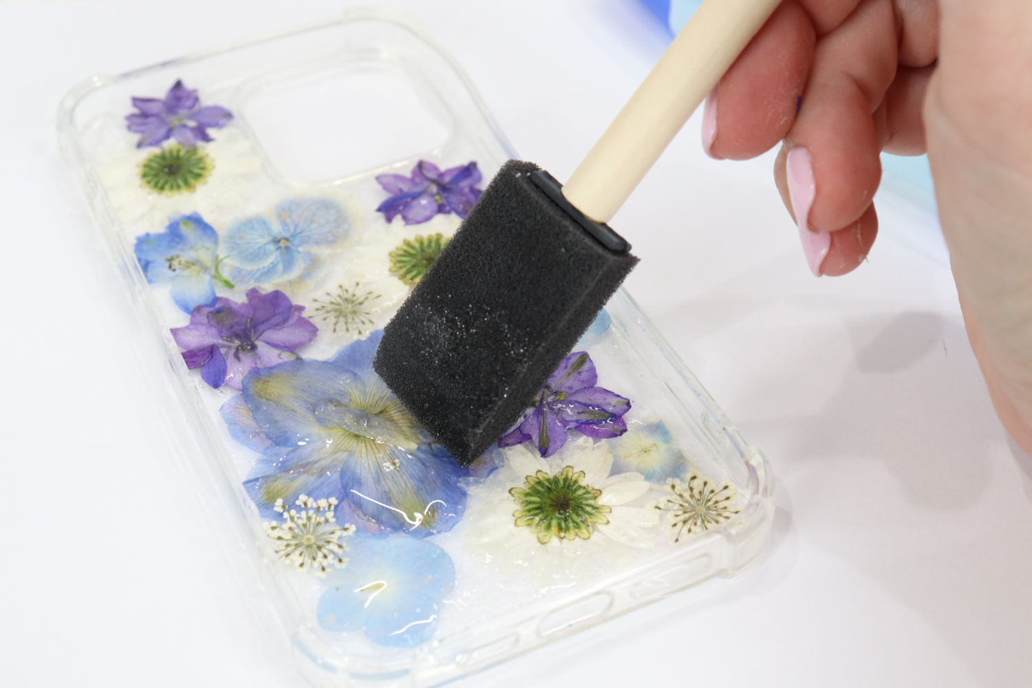 Image contains Amy’s hand using a sponge brush to apply a layer of clear glue on top of pressed flowers in a clear phone case.