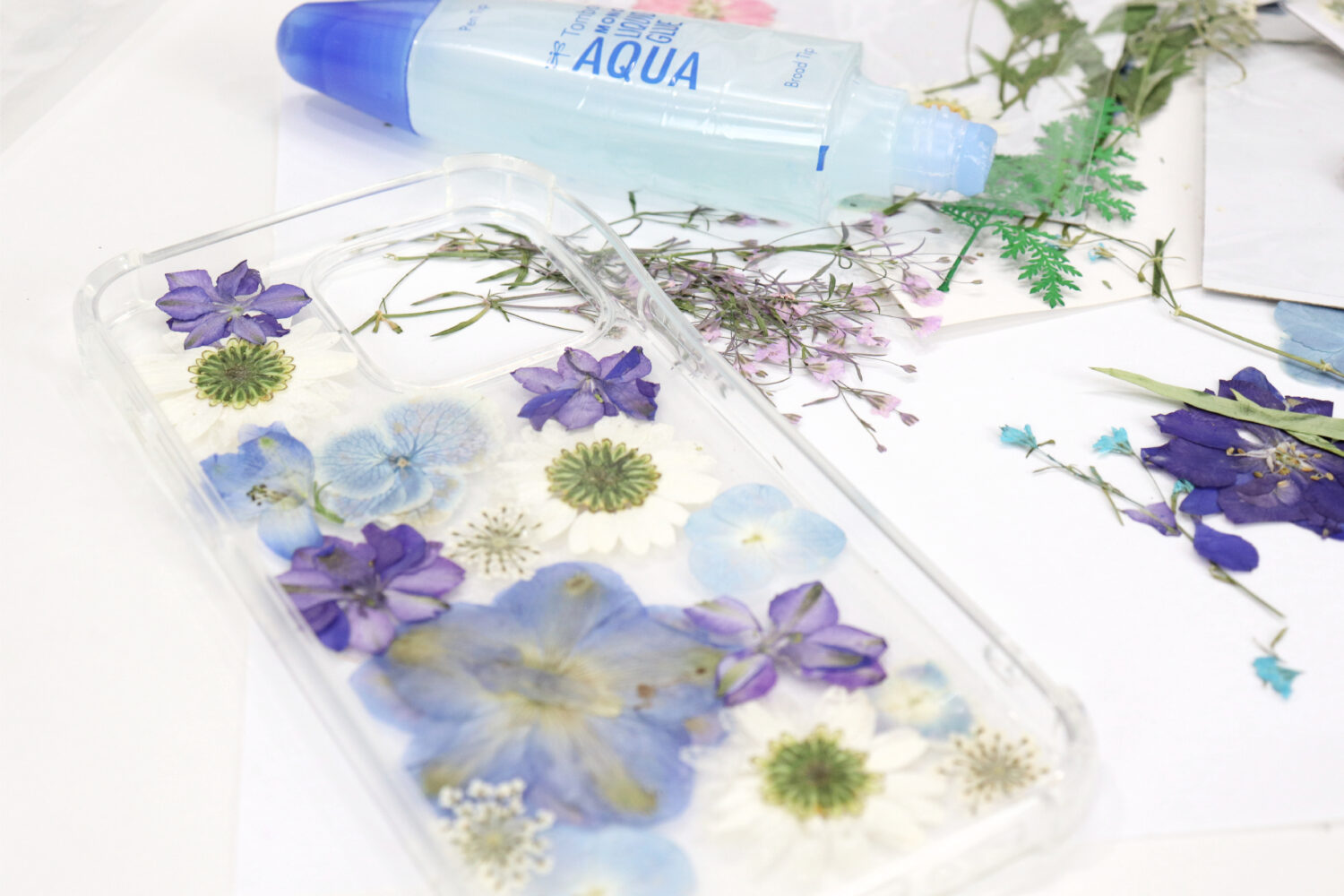 Image contains a clear phone case covered with pressed blue, purple, and white flowers. A tube of glue sits nearby, along with unused pressed flowers on a white table.