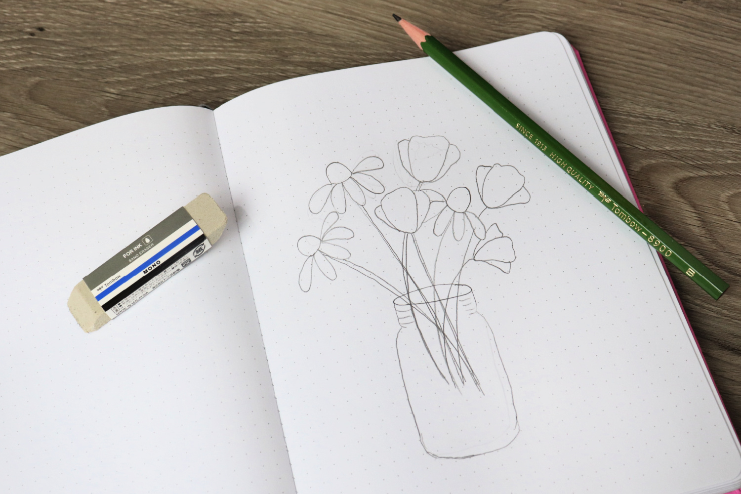 Image contains an open sketchbook with a bouquet of flowers in a mason jar sketched in pencil. A green pencil sits on top of the page, and a MONO Sand Eraser sits on the sketchbook as well.