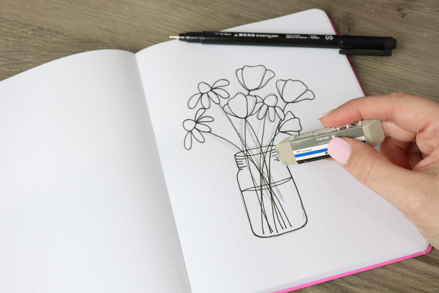 Image contains an open sketchbook with a bouquet of flowers drawn in black ink. Amy’s hand holds a MONO Sand Eraser prepared to erase any visible pencil lines. A MONO Drawing Pen 05 sits on top of the page.
