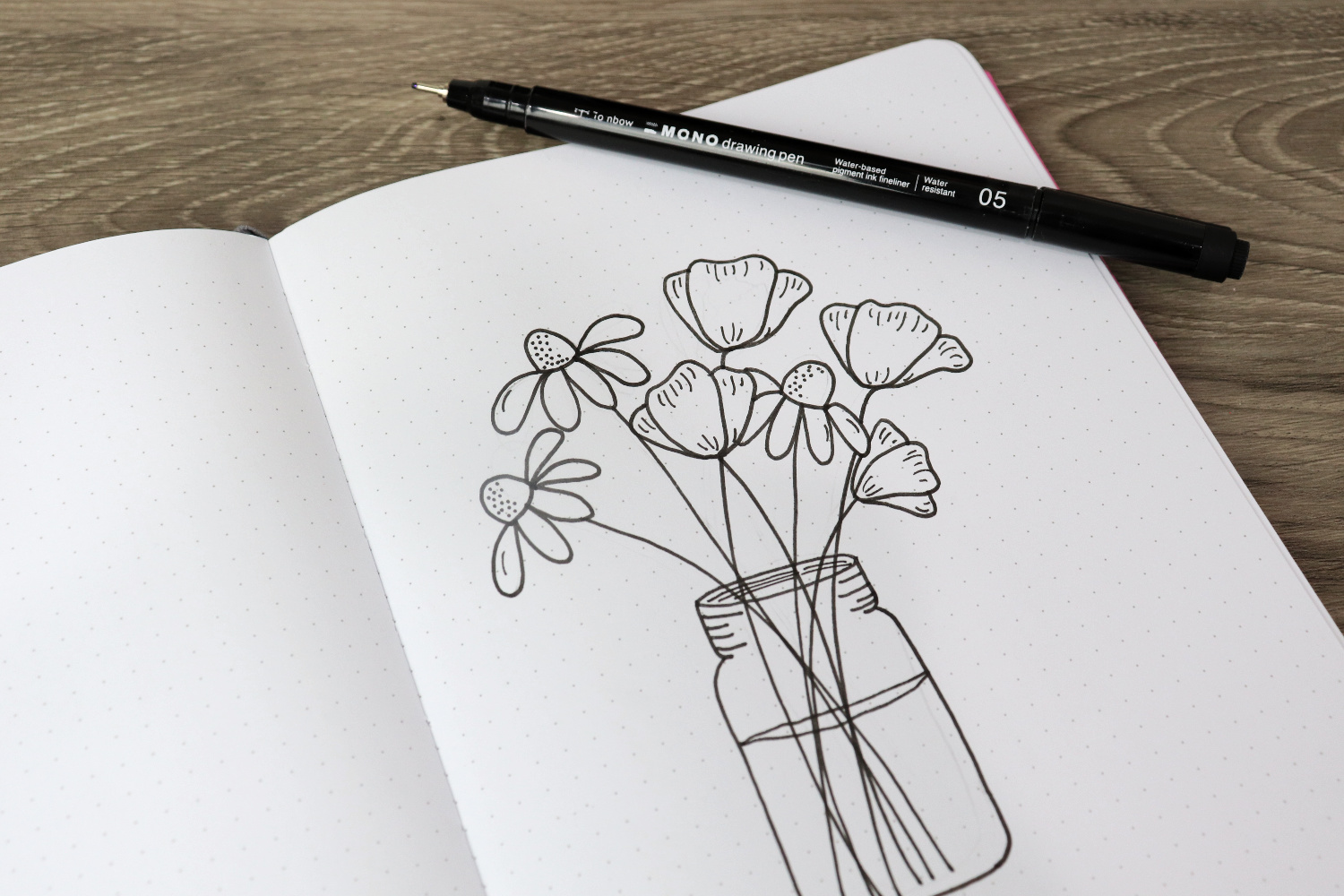 Image contains an open sketchbook with a bouquet of flowers in a mason jar drawn in black ink. A MONO Drawing Pen 05 sits on top of the page.