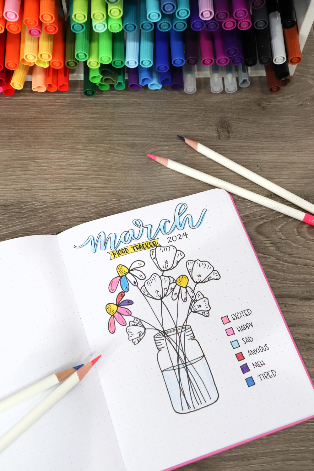 Image contains an open journal with a hand drawn flower bouquet mood tracker page. Four colored pencils sit nearby, and an organizer filled with multicolored markers is in the background on a wooden desktop.
