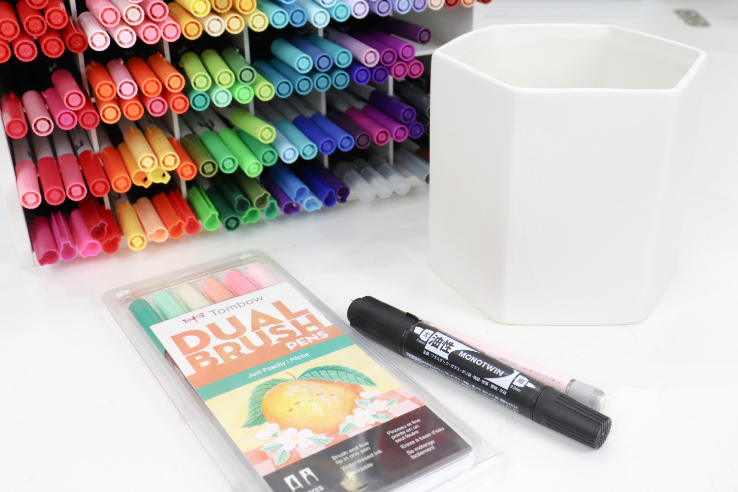 Image contains a white ceramic planter on a white table. Next to it are a pink mechanical pencil, a black permanent marker, and a pack of Dual Brush Pens. An organizer filled with rainbow colored markers sits in the background.