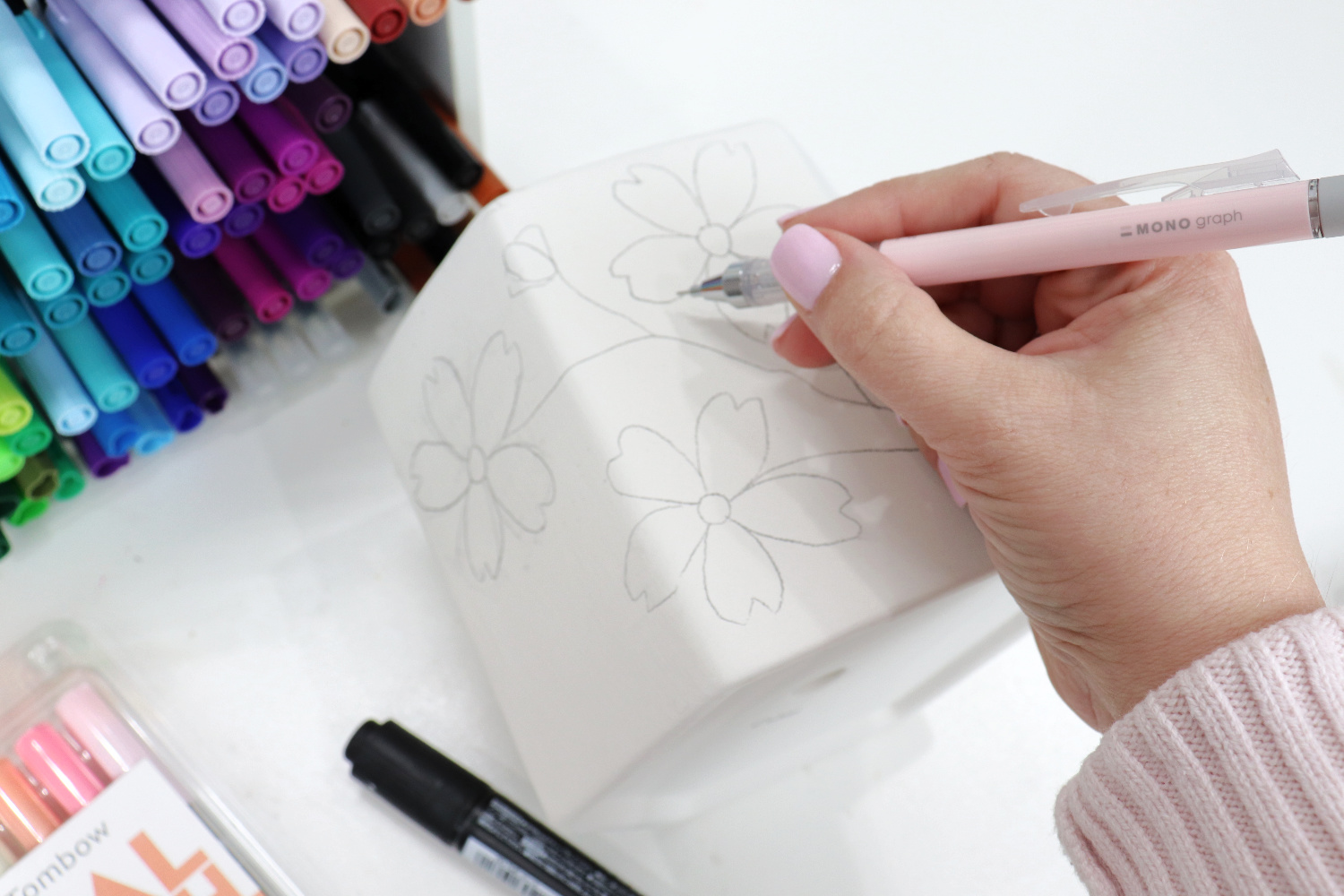 Image contains Amy’s hand holding a pink mechanical pencil and sketching cherry blossoms onto a white planter. A black marker and an organizer full of colored markers sit nearby.