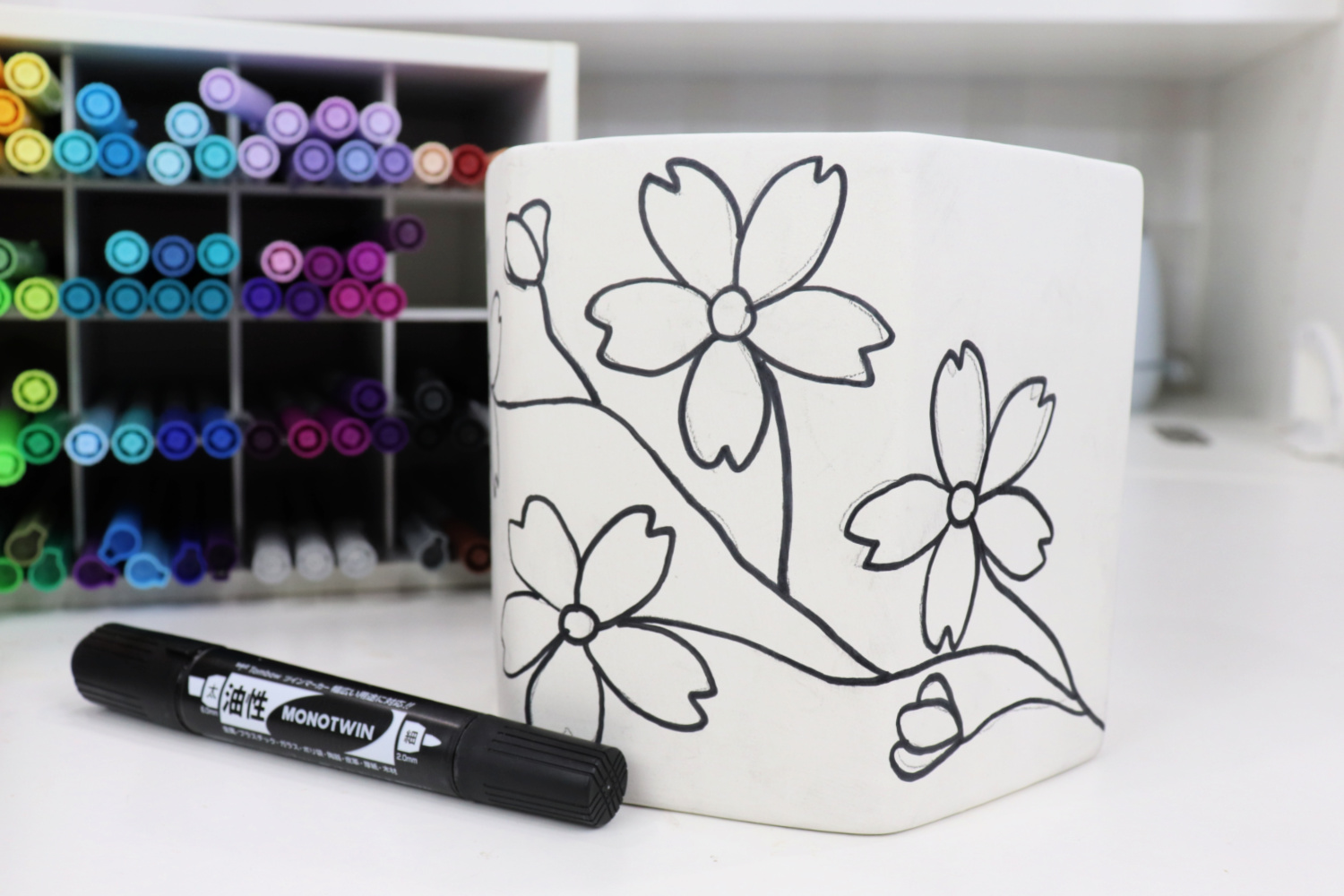 Image contains a white planter decorated with cherry blossom art outlined in black marker, sitting on a white table. A black permanent marker and an organizer full of colored markers sit nearby.