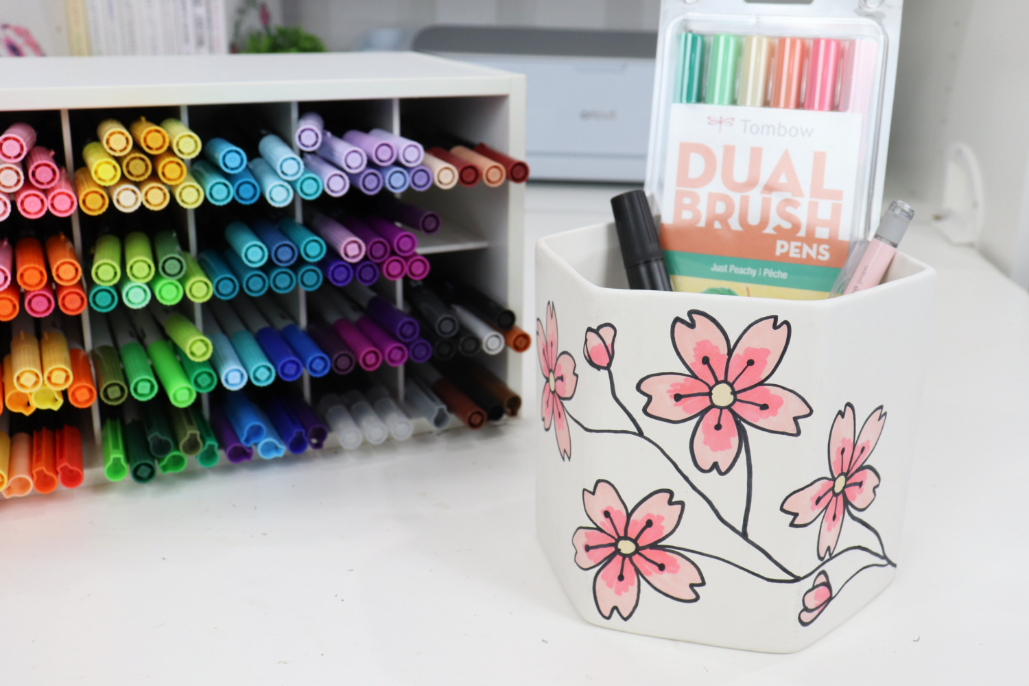Image contains the finished planter, with a pack of Dual Brush Pens, a black marker, and a pink pencil inside of it. An organizer filled with colored markers sits nearby.