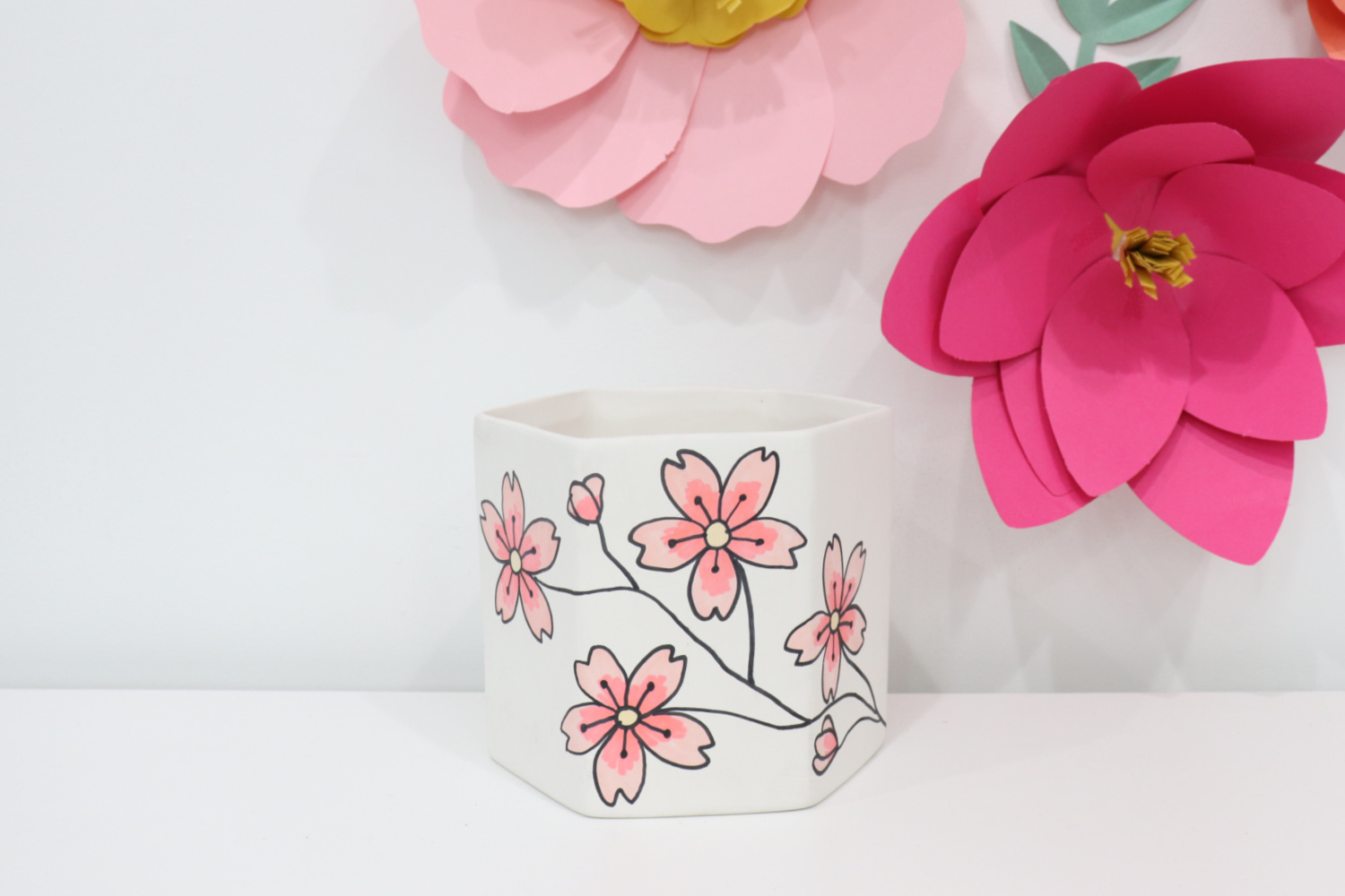 Image contains the finished cherry blossom planter sitting on a white shelf. Large light and dark. pink paper flowers hang on the wall nearby.
