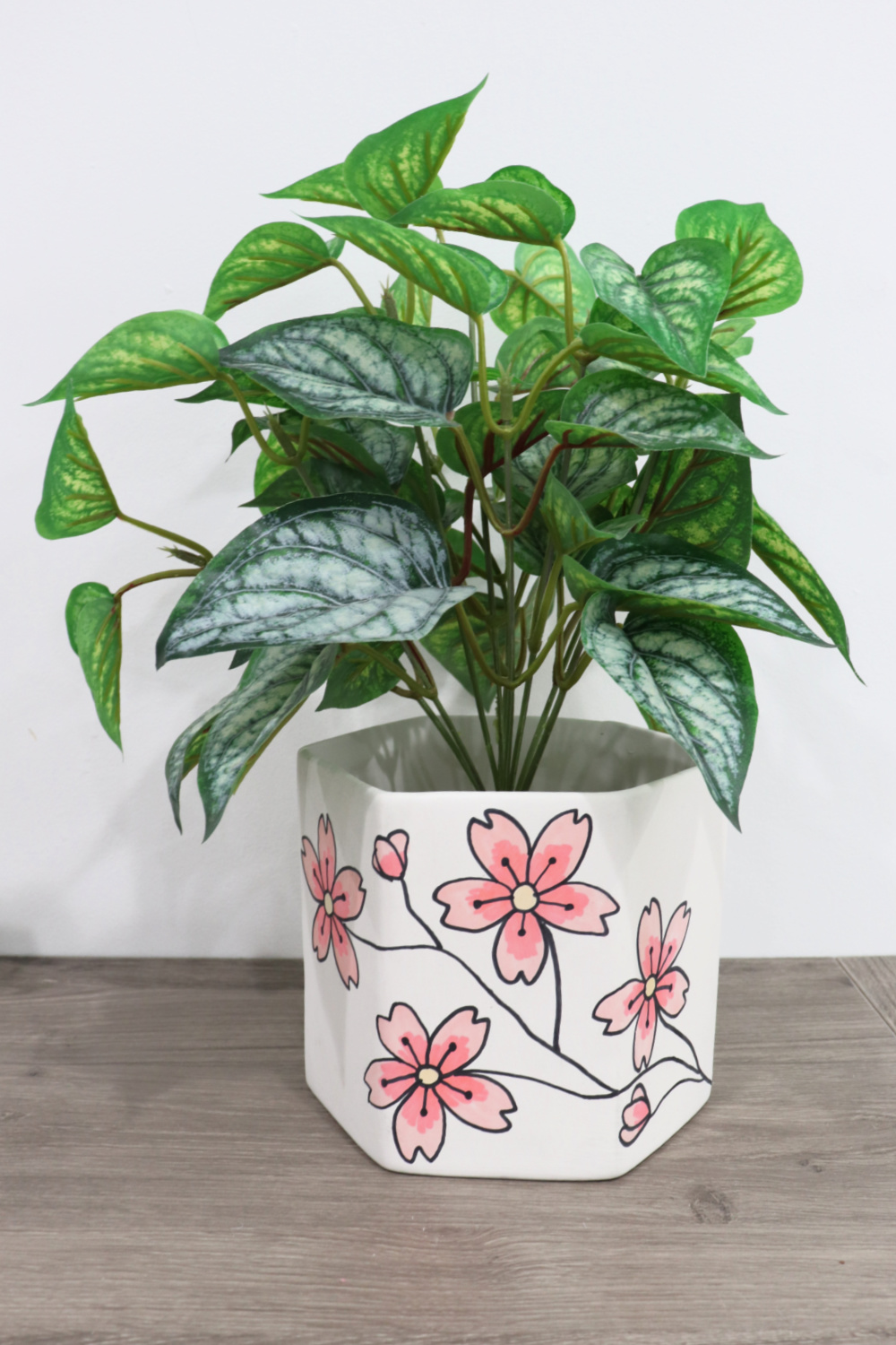 Image contains a faux plant in a white planter covered with cherry blossom illustrations. It sits on a wooden desktop.