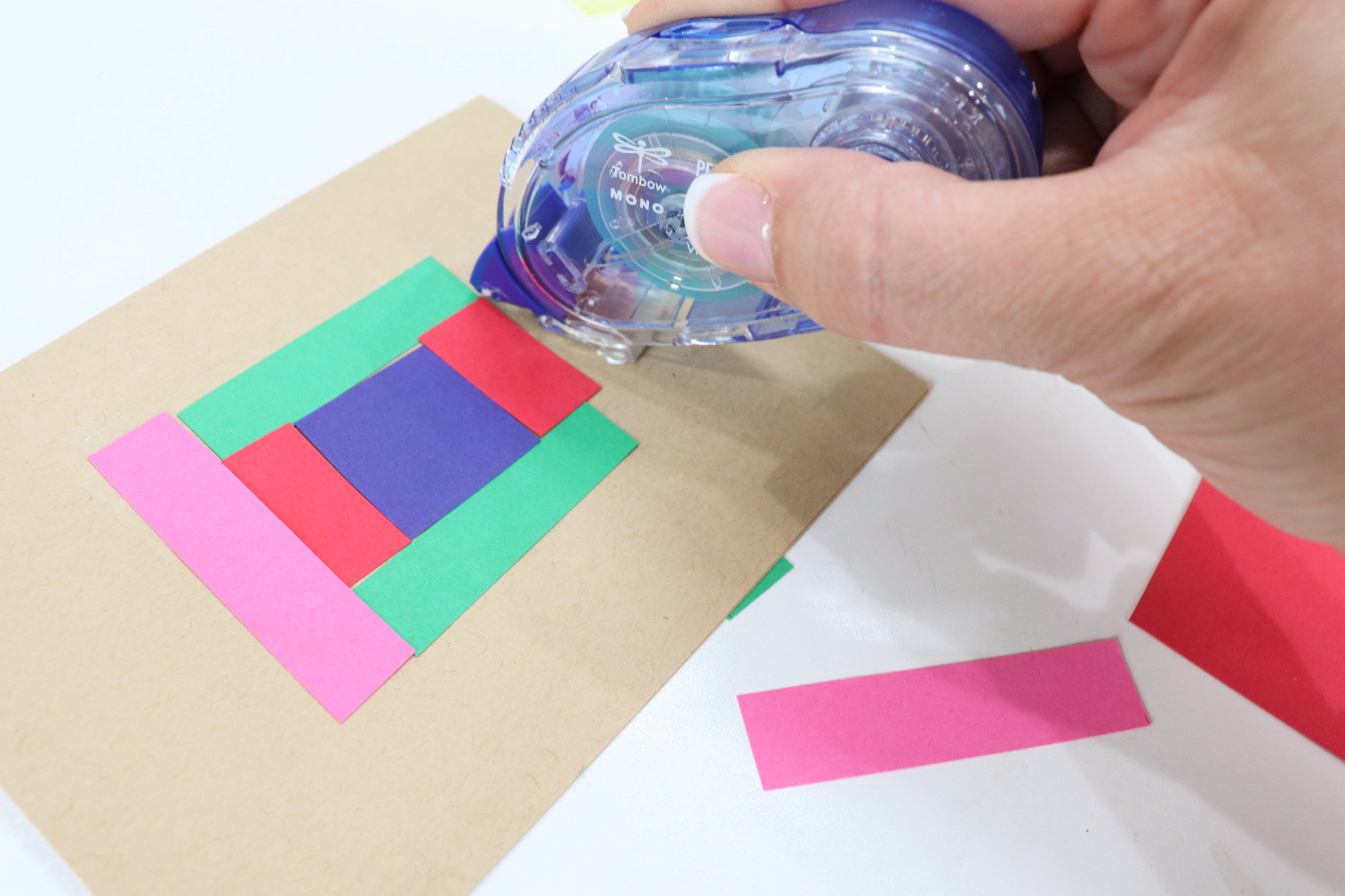 Image contains Amy’s hand holding a blue MONO Permanent Adhesive runner, applying adhesive to a kraft colored card front. Several colorful strips of cardstock are adhered to the card, which sits on a white tabletop.