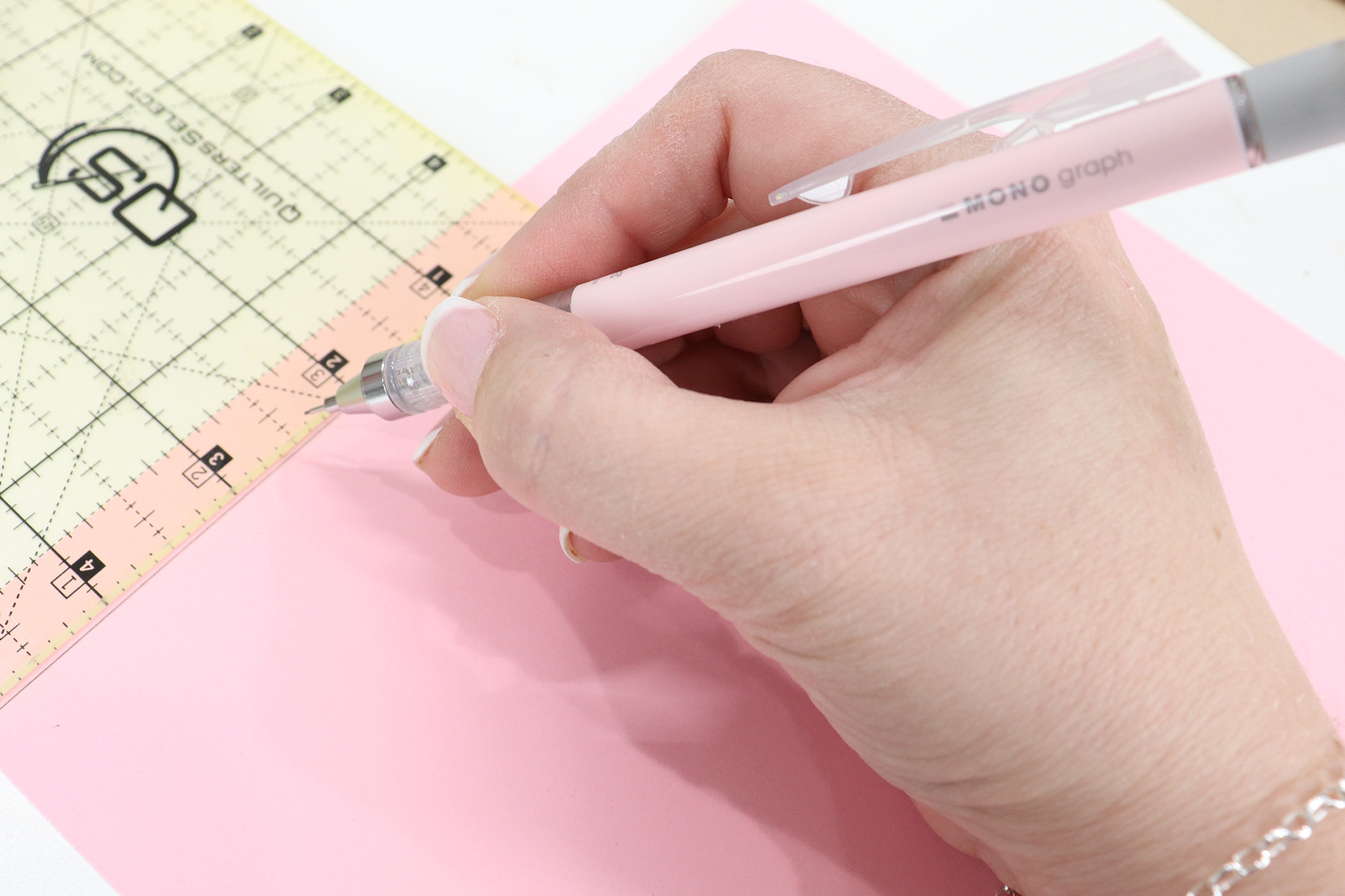 Image contains Amy’s hand holding a pastel pink MONO Graph mechanical pencil. She is drawing a line on a piece of pink cardstock, measured by a quilting ruler. 