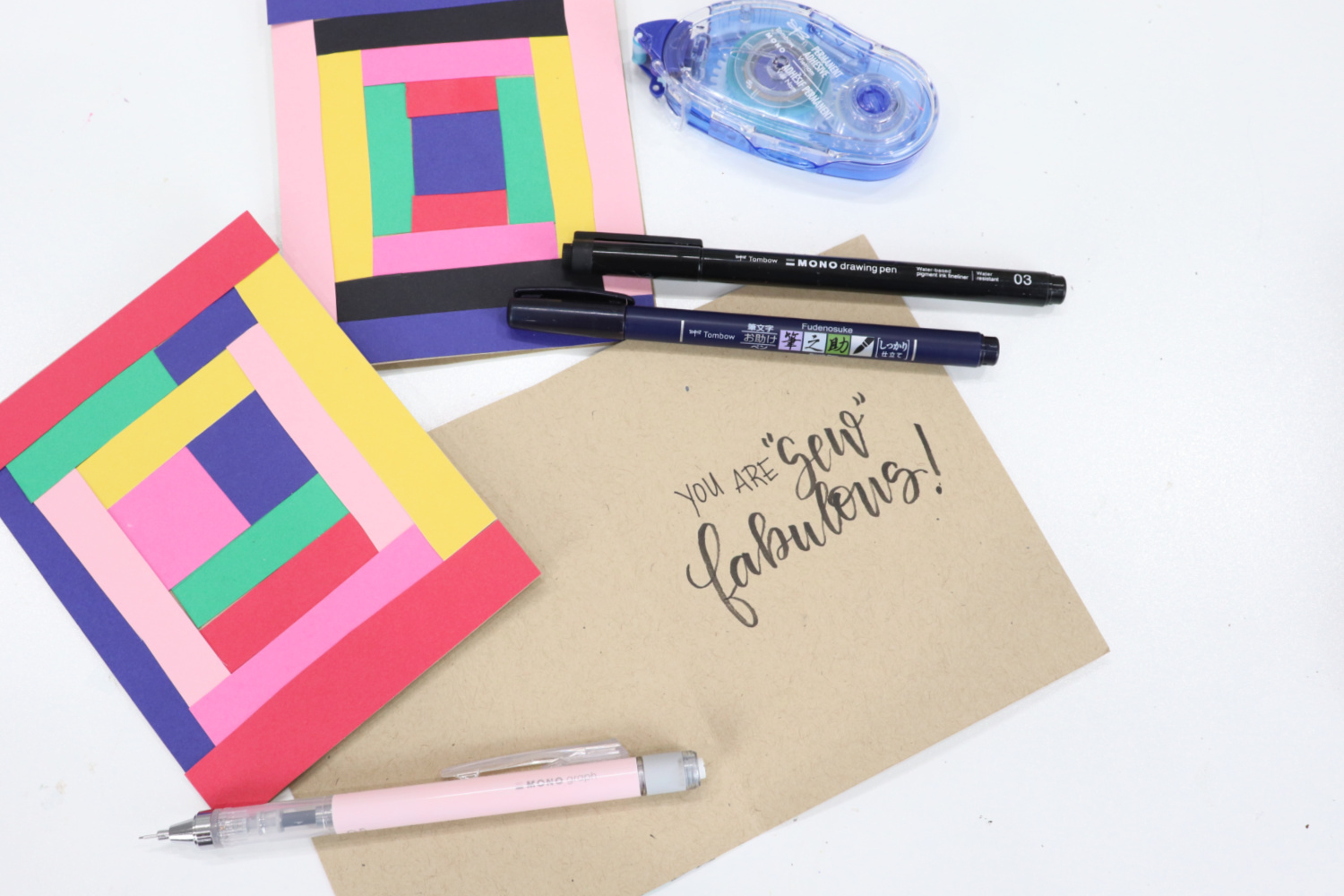 Image contains three cards; two show the fronts covered with colored cardstock strips, while the third is open to show the words, “you are ‘sew’ fabulous” written inside in black ink. Two black pens, a pink mechanical pencil, and an adhesive tape runner sit around the cards on a white table.