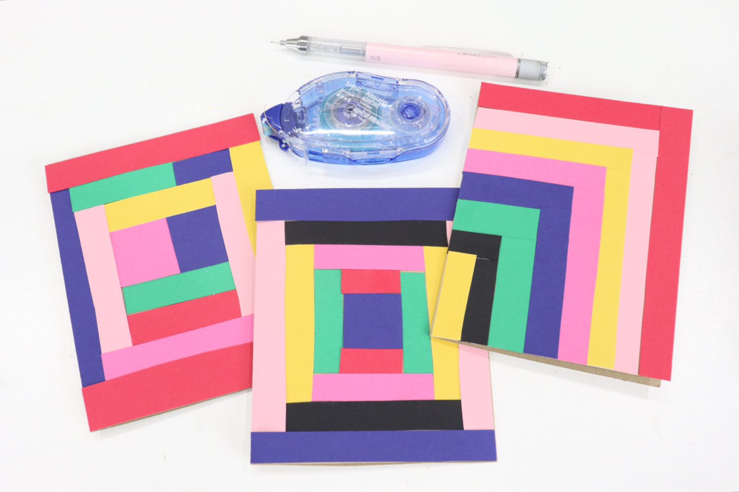 Image contains three quilt inspired cards decorated with colorful strips of cardstock. A blue permanent adhesive runner and a pink MONO Graph Mechanical Pencil sit nearby on a white tabletop.