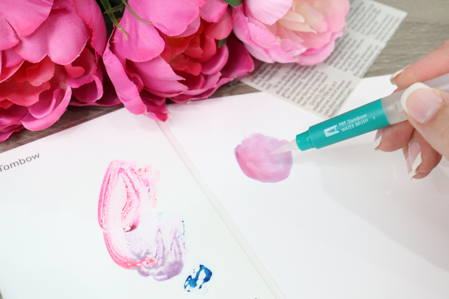 Image contains Amy’s hand holding a Water Brush and using it to paint an irregular purple shape on a piece of white Bristol board. A blending palette with blue and red ink mixed together sits in the foreground. Faux pink peonies and a newspaper clipping sit in the background.