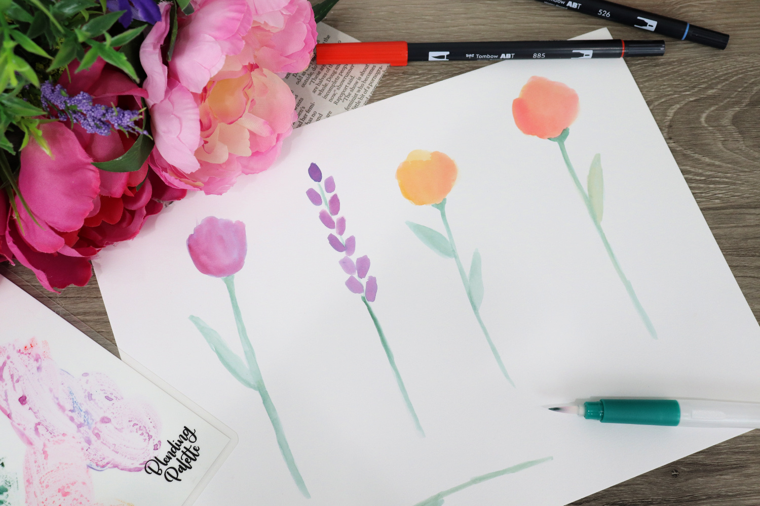 Image contains a piece of white Bristol Board that has four different flower shapes watercolored on it. Two Dual Brush Pens and a Water Pen sit nearby, along with a blending palette, faux flowers, and a newspaper clipping.