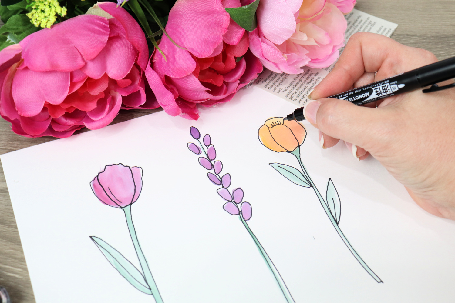 Image contains Amy’s hand holding a Tombow MONO Twin Permanent Marker and drawing a floral outline on top of the watercolored shapes. Faux flowers and a newspaper clipping sit in the background on a wooden desktop.