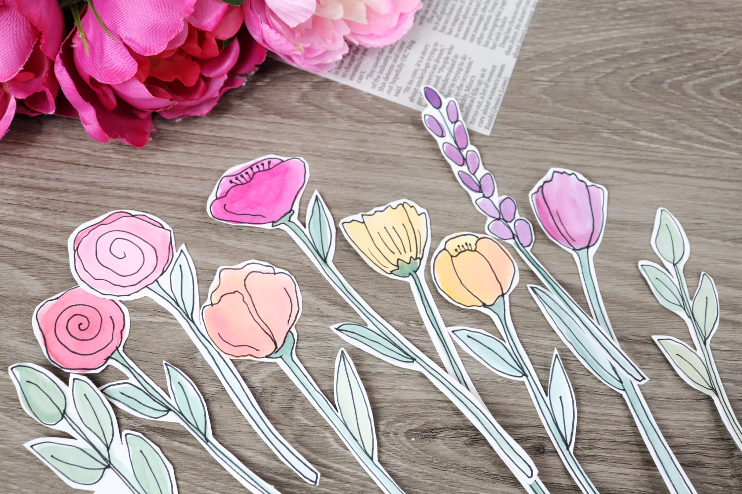 Image contains eight colorful pink, yellow, orange, and purple watercolor flowers and two leafy stems, all cut out close to the outlines. They sit on a wooden surface with faux flowers and a piece of newspaper in the background.