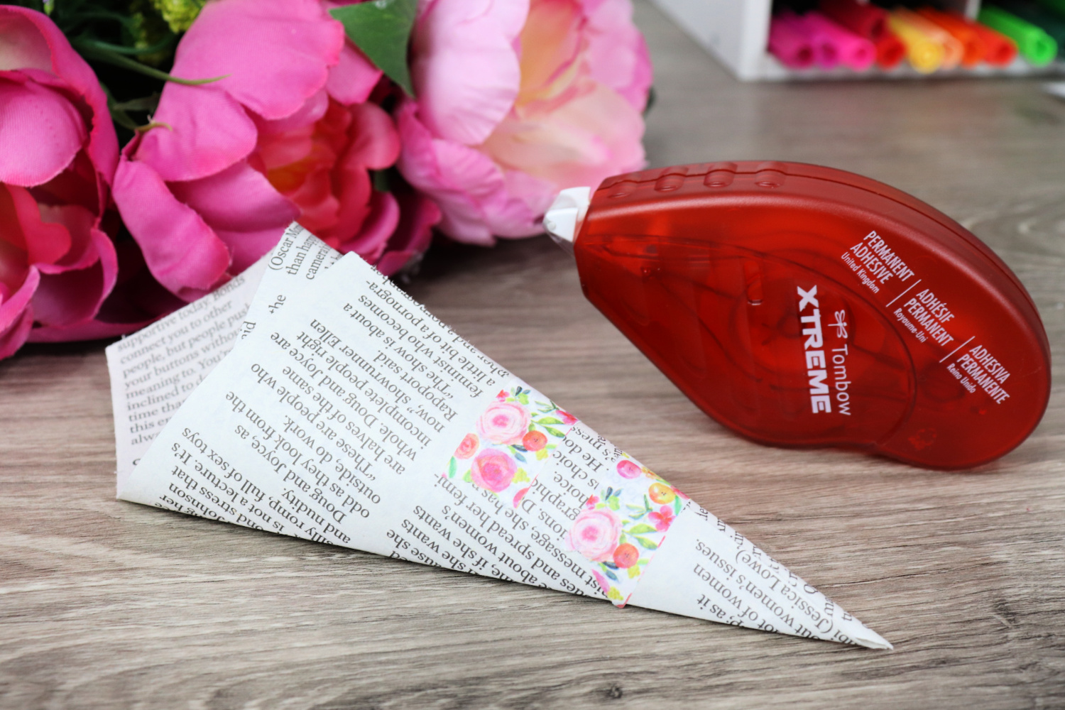 Image contains a piece of newspaper rolled into a small cone and secured with decorative pink floral washi tape. A Tombow Xtreme Tape Runner sits nearby. Three faux pink flowers and a marker caddy sit in the background on a wooden desk.