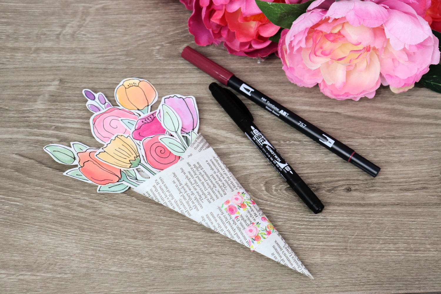Image contains a bouquet made of 10 watercolor flowers and stems tucked into a piece of wrapped newspaper. Two Tombow markers and several silk flowers sit nearby on a wooden surface.