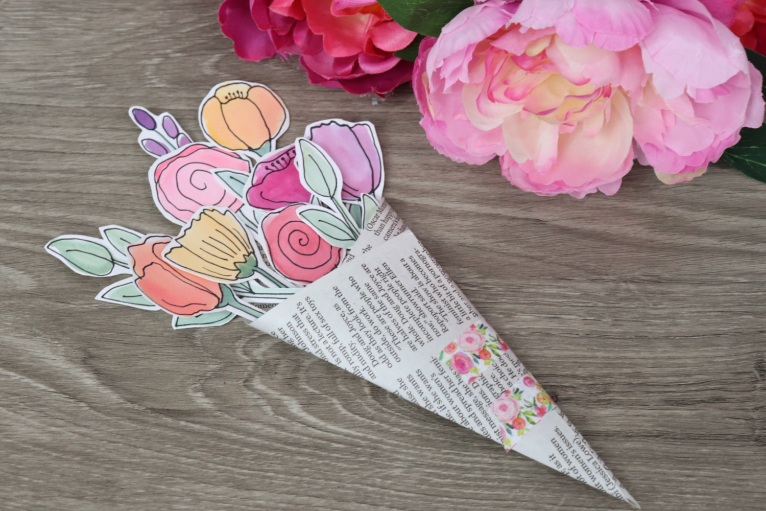 Image contains a bouquet of watercolor flowers wrapped in a newspaper clipping sitting on a wooden desk near a few pink faux flowers.