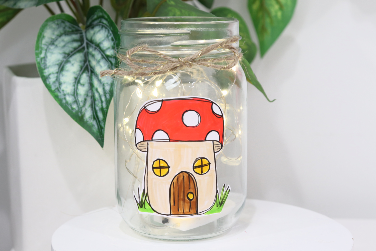 Image contains a mason jar with a toadstool house sticker on the front, filled with fairy lights. A piece of twine is tied around the mouth of the jar. It sits on a white surface with a faux plant behind it.