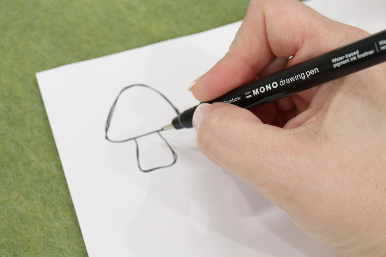 Image contains Amy’s hand holding a MONO Drawing Pen and sketching a toadstool on white printable vinyl. The background is olive green felt.
