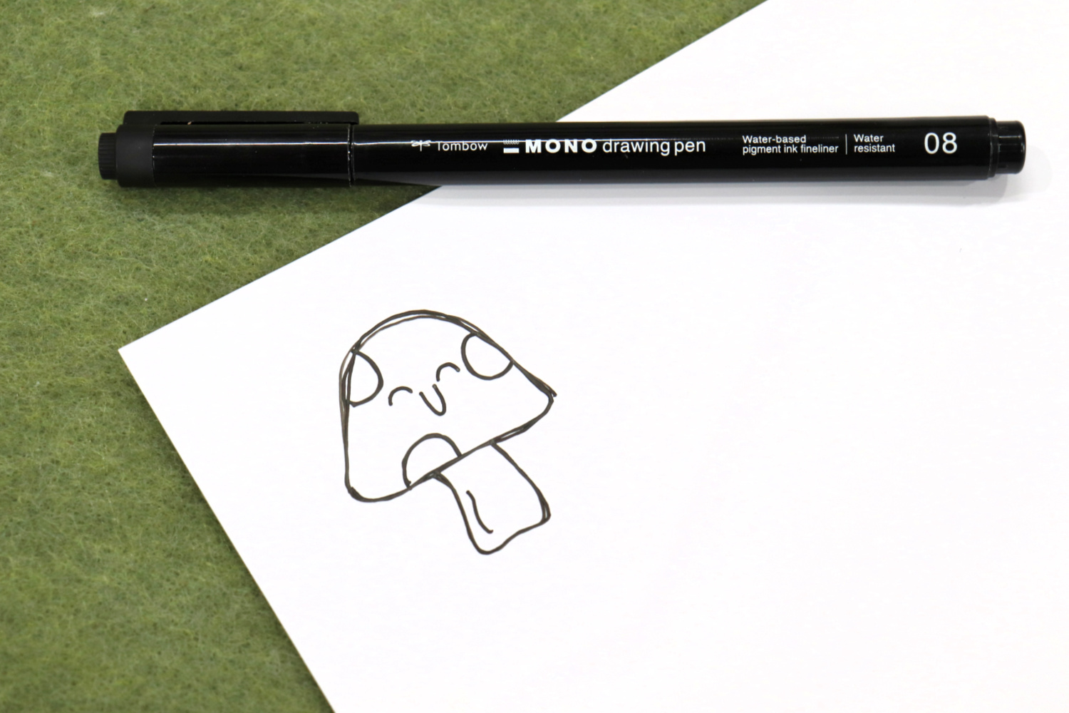 Image contains a piece of white printable vinyl with a whimsical toadstool doodled in the corner. A MONO Drawing Pen sits on top of it on a green felt background.