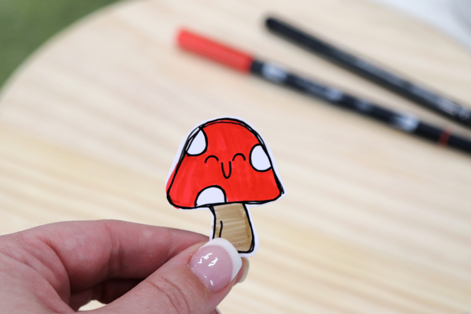 Image contains Amy’s hand holding a toadstool sticker cut from printable vinyl. In the background, two markers (red and black) sit on a wooden surface.