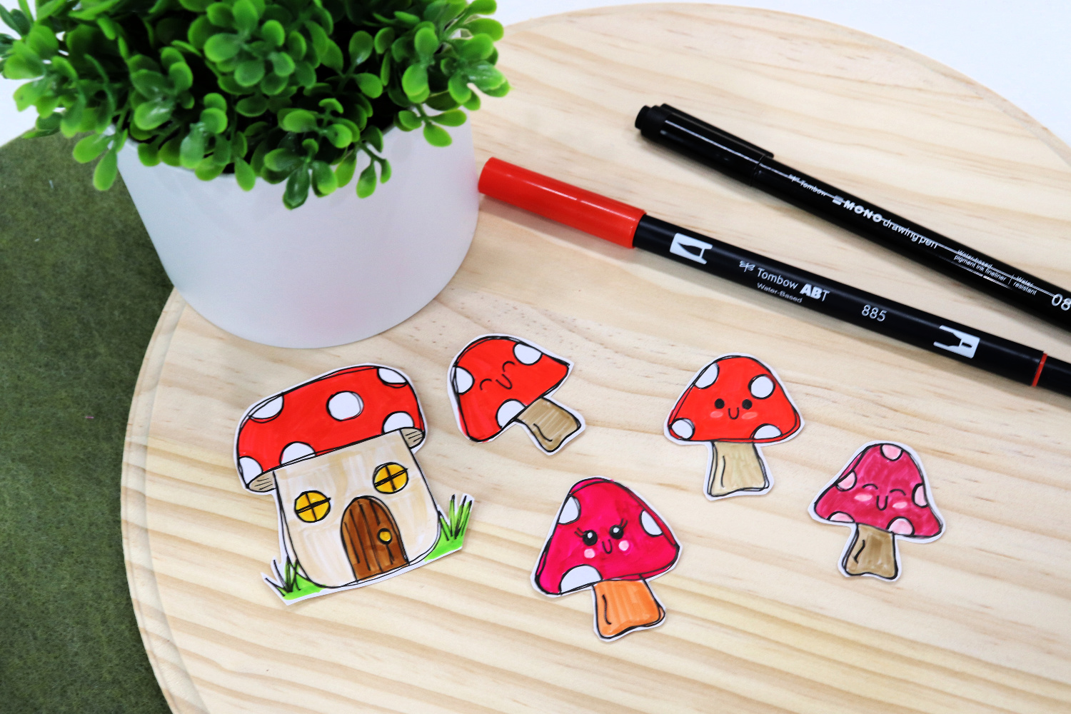Image contains hand-doodled stickers; four toadstools and a toadstool house, sitting on a wooden circle. A red and a black marker sit nearby, along with a faux plant.