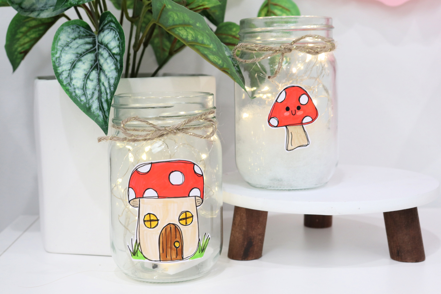 Image contains two completed cottage core lanterns; one decorated with a toadstool house, and one with a smiling toadstool. A faux plant sits in the background.