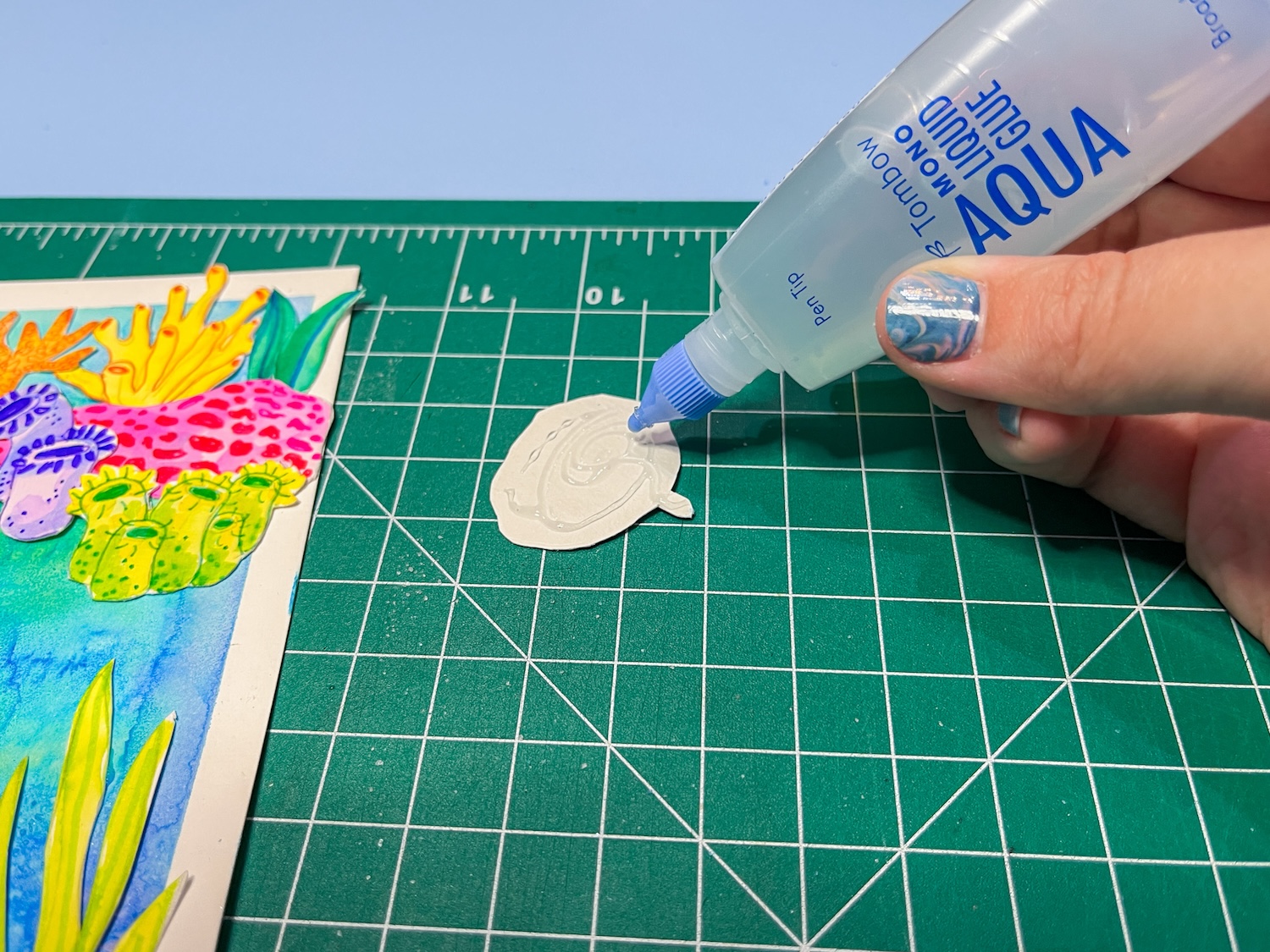 Hand holding a bottle of @tombowusa MONO Aqua Liquid Glue, applying it to the back of paper to create a DIY Ocean Collage Art