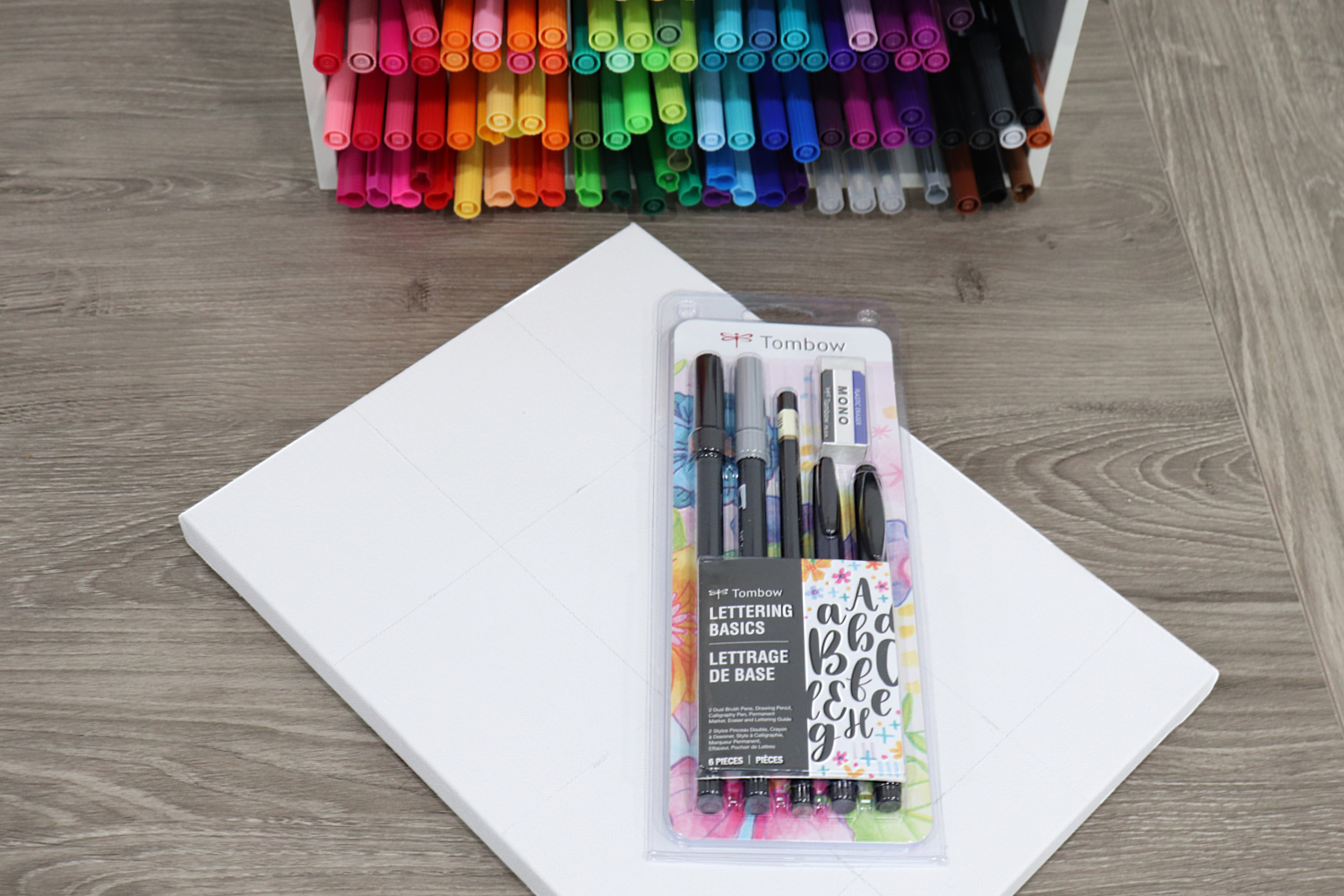 Image contains a blank white canvas sitting on a wooden desk, with the Tombow Lettering Basics Set on top of it. An organizer full of multi-colored markers sits nearby.